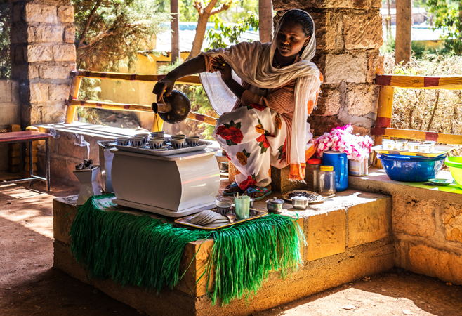 A host carefully pouring hot coffee from a black clay jebena into small, handleless cups arranged neatly on a tray